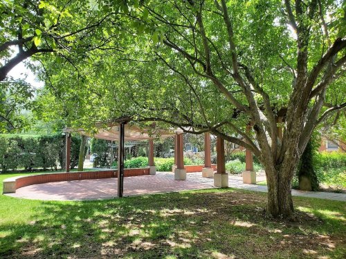 A shaded outdoor pavilion surrounded by greenery and trees, with a brick floor and stone seating.