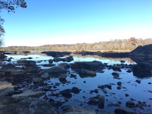 A serene river scene with smooth rocks and autumn foliage reflecting in the water under a clear blue sky.