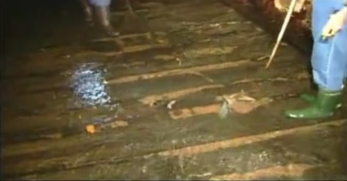 A person in green boots stands on a wet, wooden surface, with water pooling around and another figure in the background.