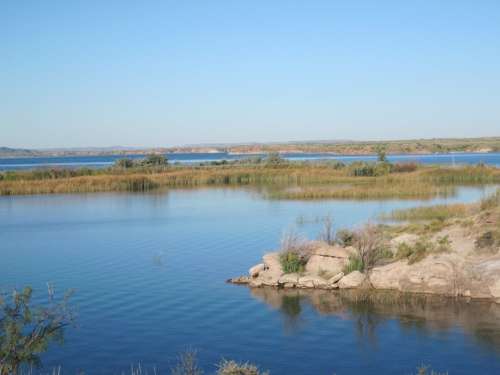 A serene lake scene with calm water, rocky shore, and lush greenery under a clear blue sky.