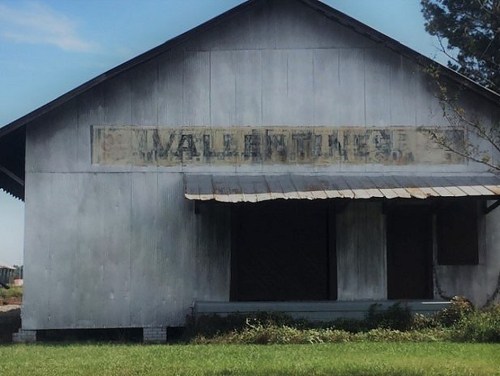 Weathered building with faded "Valentine" sign, surrounded by grass and trees under a clear blue sky.