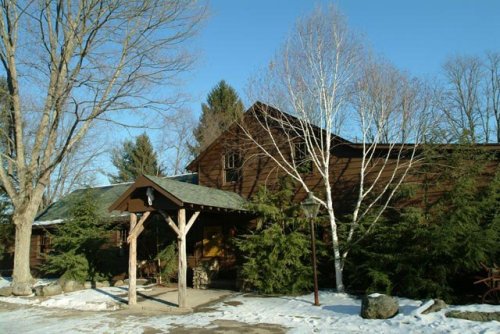 A rustic wooden cabin surrounded by trees and snow, with a clear blue sky above.