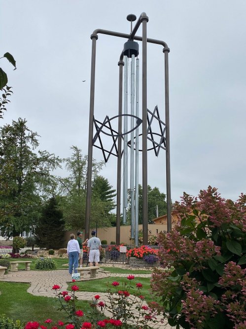 A tall metal structure with glass tubes and star motifs, surrounded by colorful flowers and two people admiring it.