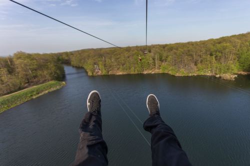 A person's feet hang above a lake while ziplining over a lush, green landscape.