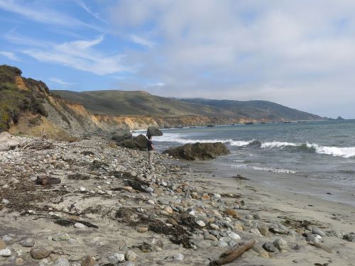 A rocky beach with gentle waves, cliffs in the background, and a person walking along the shore under a cloudy sky.