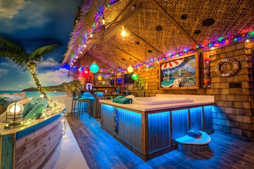 A vibrant tiki bar with colorful lights, tropical decor, and a beach view, featuring a hot tub and cozy seating.