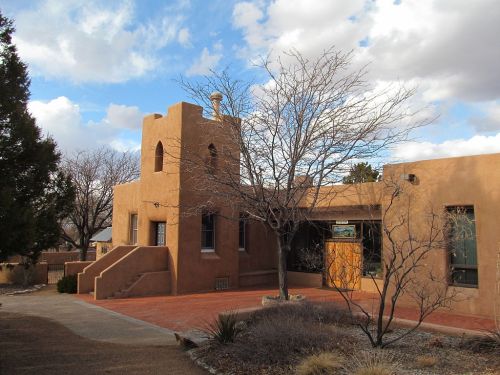 Adobe-style building with a tower, surrounded by bare trees and a blue sky with clouds. Pathway leads to the entrance.