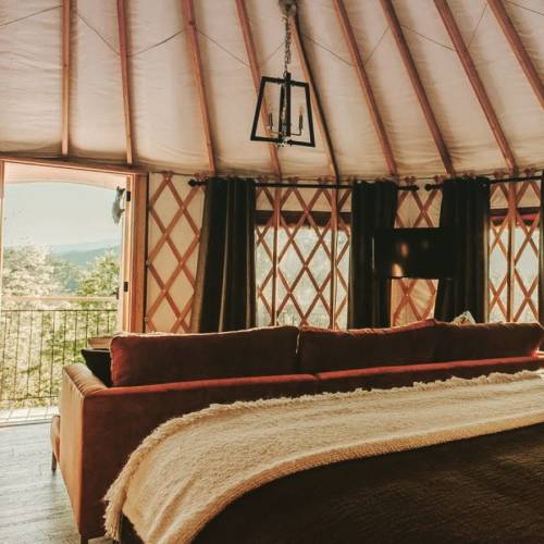 Cozy interior of a yurt with a sofa, large windows, and a view of nature, featuring warm lighting and rustic decor.
