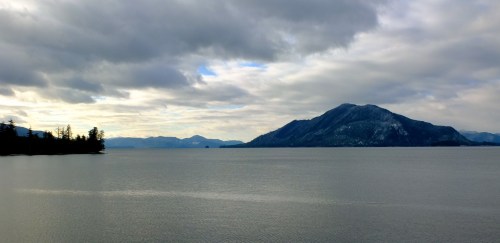 A serene lake scene with mountains in the background under a cloudy sky. Trees line the shore, reflecting on the water.