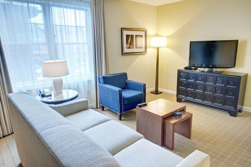 Cozy hotel room with a sofa, blue armchair, coffee table, lamp, and TV on a dresser near a window.