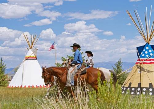 Two riders on horseback pass by colorful tipis under a blue sky with fluffy clouds and an American flag in the background.