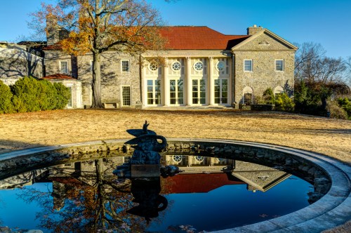 A stone building with large windows reflects in a pond, surrounded by autumn trees and a clear blue sky.