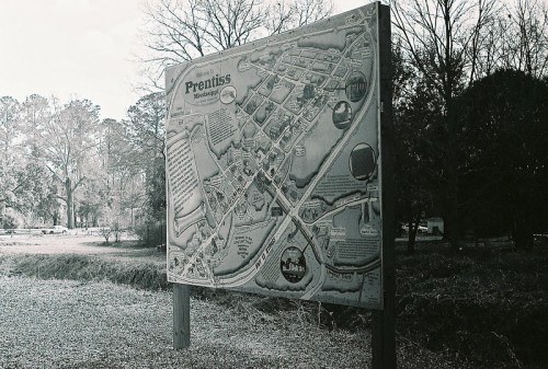 Map of Prentiss, Mississippi, displayed on a sign, surrounded by trees and a gravel area. Black and white photo.