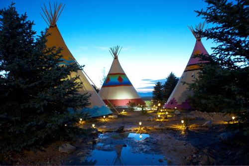 Three colorful tipis illuminated at dusk, surrounded by trees and reflecting in a small puddle.