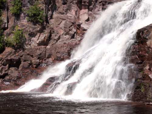 A cascading waterfall flows over rocky cliffs, surrounded by lush greenery and reflecting in the water below.