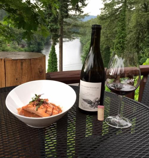 A plate of salmon with a wine bottle and glass, set against a scenic view of trees and a lake.