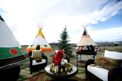 Three colorful tipis surround a cozy seating area with a table, decorated with blankets and drinks, against a cloudy sky.