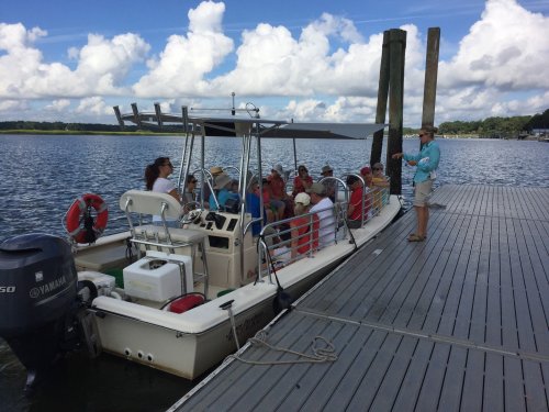 A group of people on a boat at a dock, with a guide speaking to them by the water under a cloudy sky.