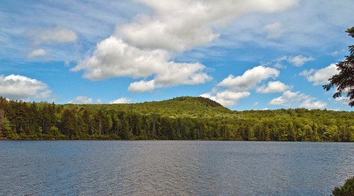 A serene lake surrounded by lush green trees and a hill under a partly cloudy blue sky.