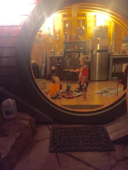 Two toddlers play inside a cozy, circular room with wooden walls and a kitchen visible in the background.