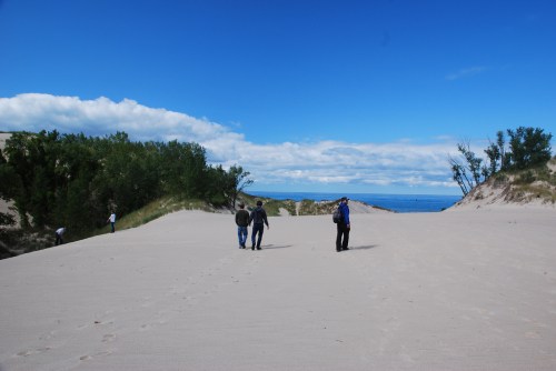 Two people walk along a sandy path with trees and a blue sky in the background, near a body of water.
