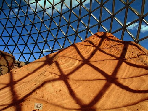 A sandy mound with geometric shadows cast by a glass dome against a blue sky.