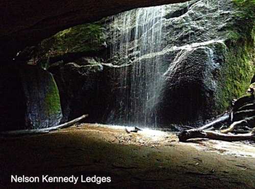 A serene waterfall cascades over moss-covered rocks in a cave at Nelson Kennedy Ledges, with soft light illuminating the scene.