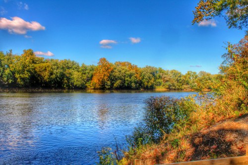 A serene river scene with lush green trees and autumn foliage under a clear blue sky.