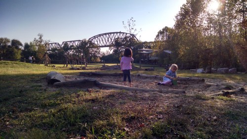 Two children play in a sandy area at a park, with a bridge and trees in the background under a clear sky.