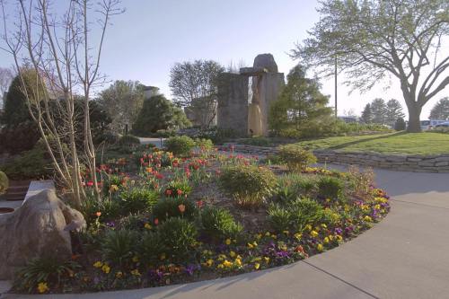 A landscaped garden featuring colorful flowers, stone structures, and lush greenery under a clear blue sky.