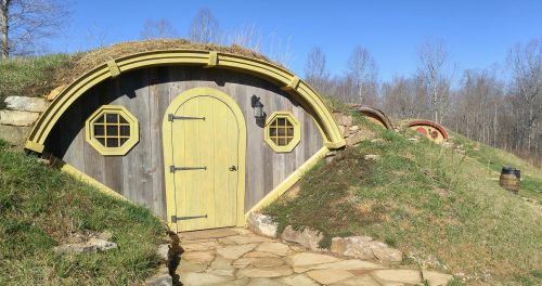 A whimsical hobbit-style house with a round door and windows, nestled in a grassy landscape under a clear blue sky.