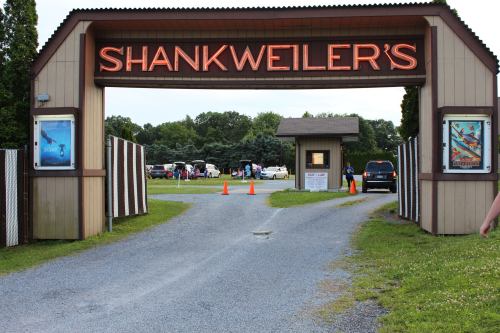 Entrance to Shankweilers, featuring a neon sign and a gravel path leading into a grassy area with people and vehicles.