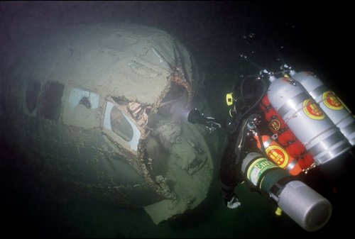 A diver explores a sunken airplane wreck, partially covered in sediment, underwater in a dark environment.