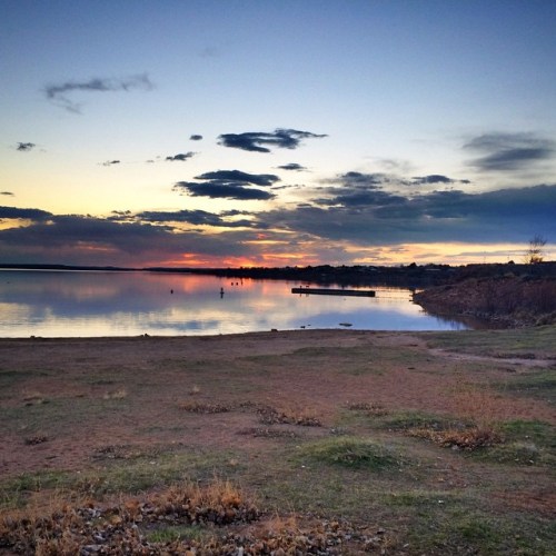 A serene lakeside view at sunset, with calm waters reflecting colorful clouds and a distant shoreline.