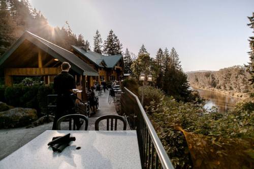 A scenic restaurant patio overlooking a river, surrounded by trees, with a waiter preparing for guests.