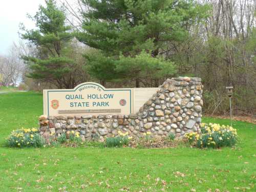 Welcome sign for Quail Hollow State Park, featuring stonework and blooming flowers in a green landscape.