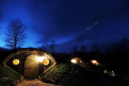 A cozy hobbit-style home illuminated at night, surrounded by trees under a dramatic blue sky.