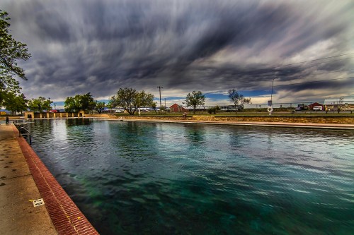 A calm river reflects a dramatic sky with dark clouds, lined by trees and buildings in the background.