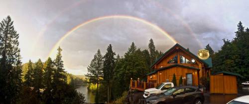 A scenic view of a wooden cabin by a lake, framed by tall trees and a vibrant rainbow in the sky.