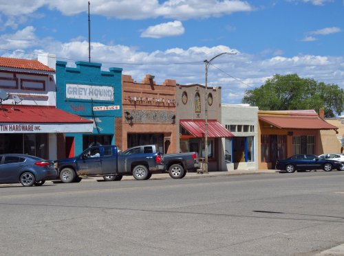 Colorful storefronts line a street, featuring a mix of businesses and parked vehicles under a blue sky with clouds.