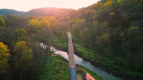 Aerial view of a wooden bridge over a winding river, surrounded by lush trees and autumn foliage at sunset.