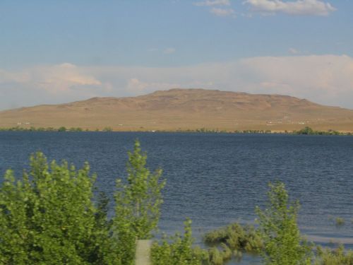 A serene lake with green foliage in the foreground and a distant, flat-topped mountain under a blue sky.