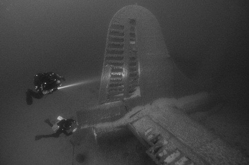 Two divers explore the wreck of an aircraft submerged underwater, illuminated by their flashlights.