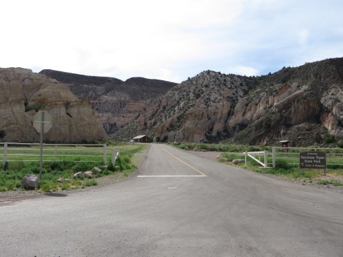 A gravel road leads into a scenic area with rocky cliffs and green grass, marked by a sign for a state park.