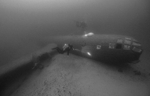 Two divers explore a sunken aircraft on the ocean floor, surrounded by murky water and marine debris.