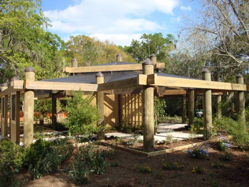 A wooden pavilion with a sloped roof surrounded by greenery and flowers under a blue sky.