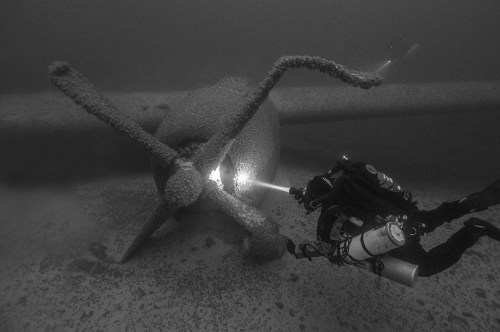 A diver explores a submerged propeller, illuminating it with a flashlight in a murky underwater environment.