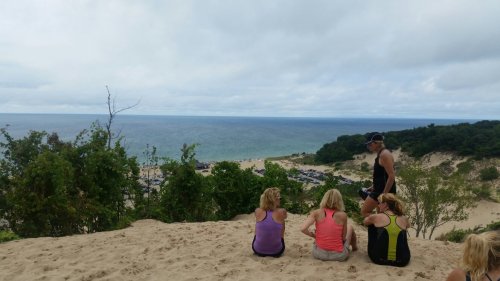 A group of people sitting on a sandy dune, overlooking a beach and ocean under a cloudy sky.