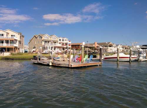 A scenic waterfront view featuring colorful chairs on a dock, with houses and boats in the background under a blue sky.