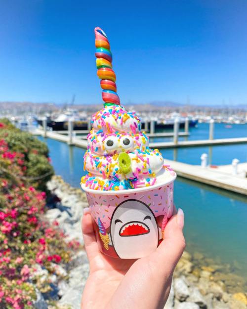 A colorful ice cream cone topped with sprinkles and a rainbow swirl, held by a hand near a marina.